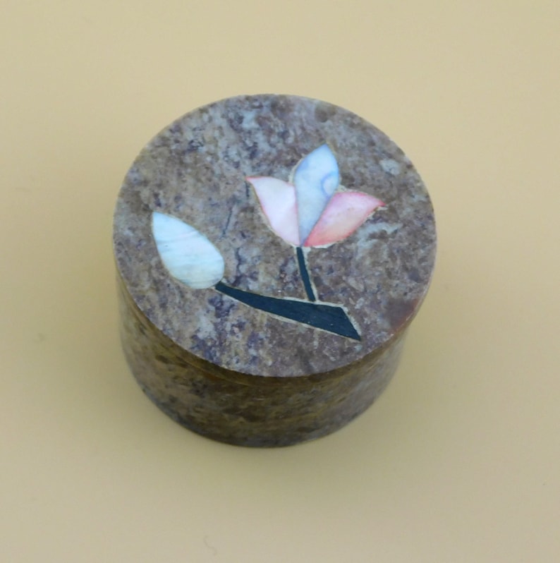 May include: A round, brown stone box with a floral design. The flower is made of inlaid pieces of colorful stone, including pink, blue, and white.