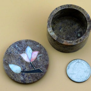 May include: A small, round, brown stone trinket box with a lid. The lid has a decorative inlay of a flower with pink, white, and blue petals.
