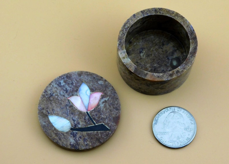 May include: A small, round, brown stone trinket box with a lid. The lid has a decorative inlay of a flower with pink, white, and blue petals.