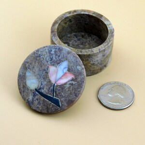 May include: A small, round, stone trinket box with a floral design. The lid is open and the inside of the box is visible. The box is made of a light brown stone with a marbled pattern. The floral design is made of inlaid pieces of different colored stones, including pink, blue, and white. The box is about the size of a quarter.