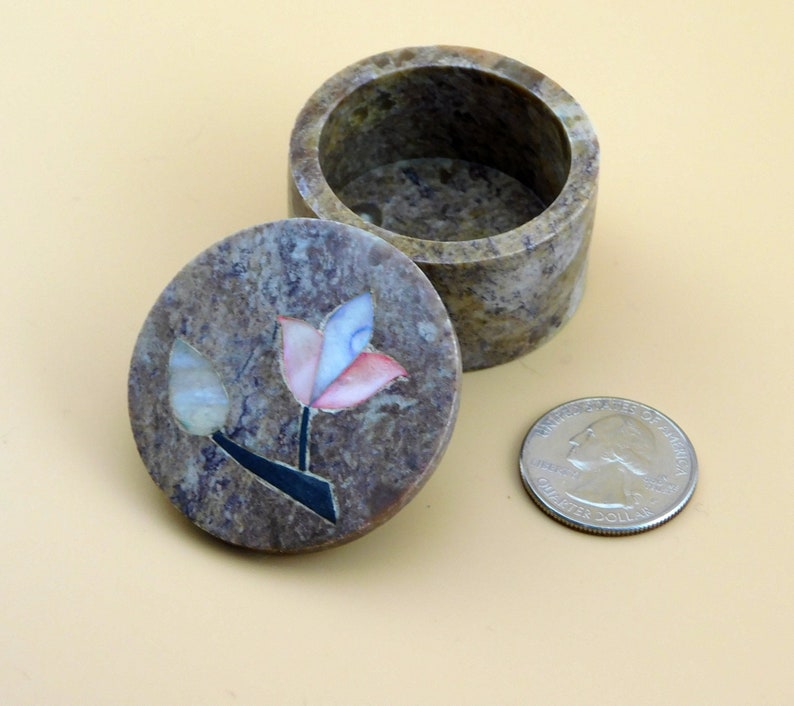 May include: A small, round, stone trinket box with a floral design. The lid is open and the inside of the box is visible. The box is made of a light brown stone with a marbled pattern. The floral design is made of inlaid pieces of different colored stones, including pink, blue, and white. The box is about the size of a quarter.