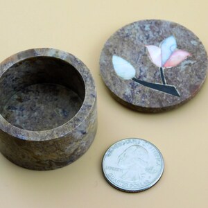 May include: A small, round, brown stone trinket box with a floral design on the lid. The lid is open and the inside of the box is visible. The box is about the size of a US quarter.