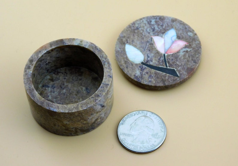 May include: A small, round, brown stone trinket box with a floral design on the lid. The lid is open and the inside of the box is visible. The box is about the size of a US quarter.