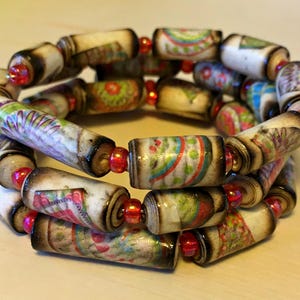 May include: A multi-layered bracelet made of colorful, patterned wooden beads. The beads are arranged in a circular pattern and feature floral and geometric designs. The bracelet is accented with small red beads.