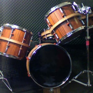 Puede incluir: Un set de batería de cuatro piezas con un bombo negro, dos toms de madera marrón y un tambor de madera marrón. Los tambores están montados en soportes con soportes de platillos en el fondo.