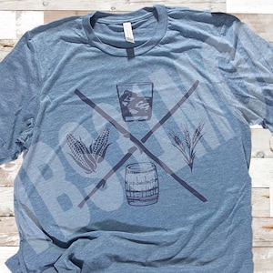 May include: Blue t-shirt with a graphic design featuring a crossed-out design with a glass of whiskey, corn, wheat, and a barrel. The shirt is made of a soft, heathered fabric.