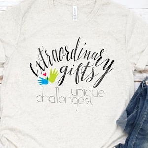 May include: A light beige t-shirt with the words "Extraordinary Gifts, Unique Challenges" in black script. The design includes a colorful hand graphic. The shirt is paired with blue jeans and white canvas sneakers.