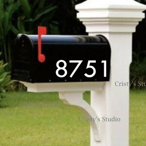 May include: A black mailbox with the number 8751 in white, mounted on a white post. The mailbox has a red flag. The post is supported by a white bracket. Green grass and foliage are in the background.