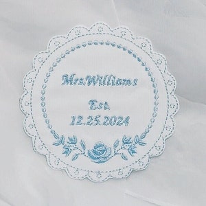 May include: A white, circular embroidered patch with a scalloped edge. The patch features light blue text that reads "Mrs. Williams Est 12.25.2024" within a floral border. The design is set against a white background.