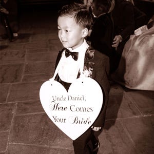 May include: A young boy in a tuxedo holds a heart-shaped sign that reads "Uncle Daniel, Here Comes Your Bride".