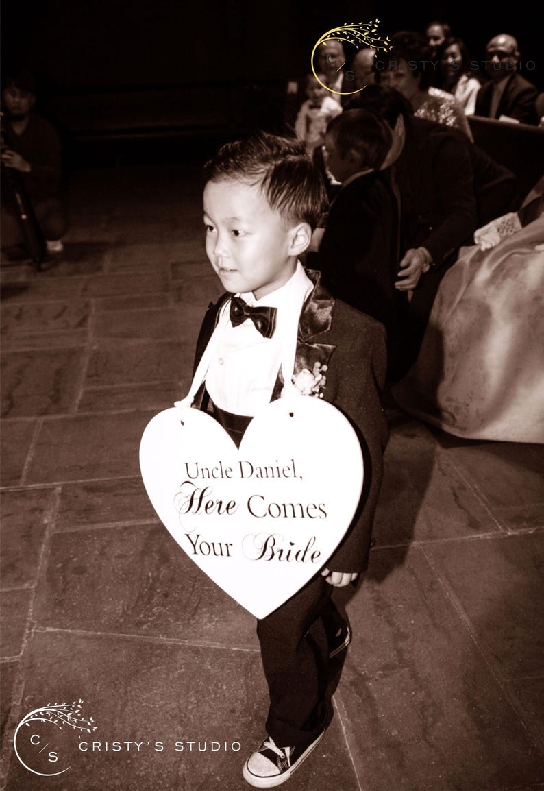 May include: A young boy in a tuxedo holds a heart-shaped sign that reads "Uncle Daniel, Here Comes Your Bride".