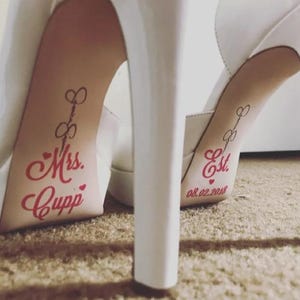 Op de afbeelding: Witte trouwschoenen met hoge hakken en rode tekst op de binnenzolen. E&eacute;n binnenzool leest "Mrs. Cupp" en de andere "Est. 08.02.2018". De schoenen hebben een plateauzool en een stilettohak.