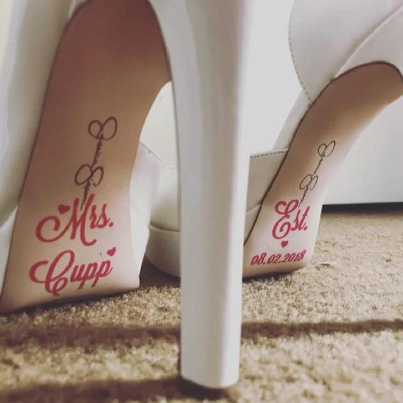 Op de afbeelding: Witte trouwschoenen met hoge hakken en rode tekst op de binnenzolen. E&eacute;n binnenzool leest "Mrs. Cupp" en de andere "Est. 08.02.2018". De schoenen hebben een plateauzool en een stilettohak.