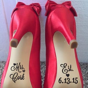 Op de afbeelding: Een paar felrode satijnen pumps met strikaccenten. De zolen zijn voorzien van de woorden "Mrs. Cork" en "Est. 6.13.15". De hakken hebben een stiletto-ontwerp.
