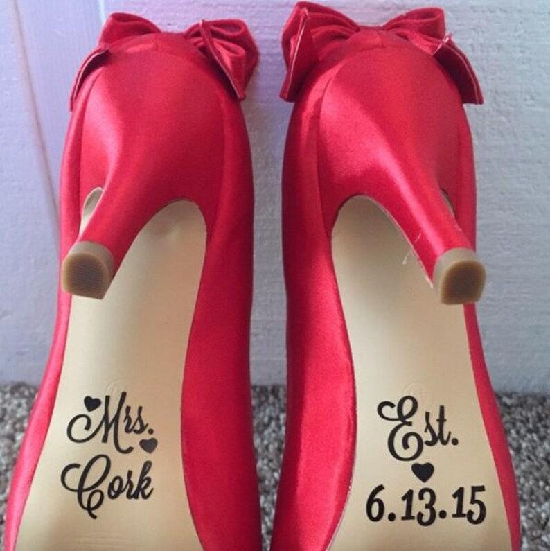 Op de afbeelding: Een paar felrode satijnen pumps met strikaccenten. De zolen zijn voorzien van de woorden "Mrs. Cork" en "Est. 6.13.15". De hakken hebben een stiletto-ontwerp.