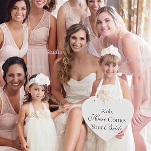 May include: A group of bridesmaids in pink dresses stand with the bride in a white wedding dress. A flower girl in a white tulle dress holds a heart-shaped sign that reads "Rob, Here Comes Your Bride".