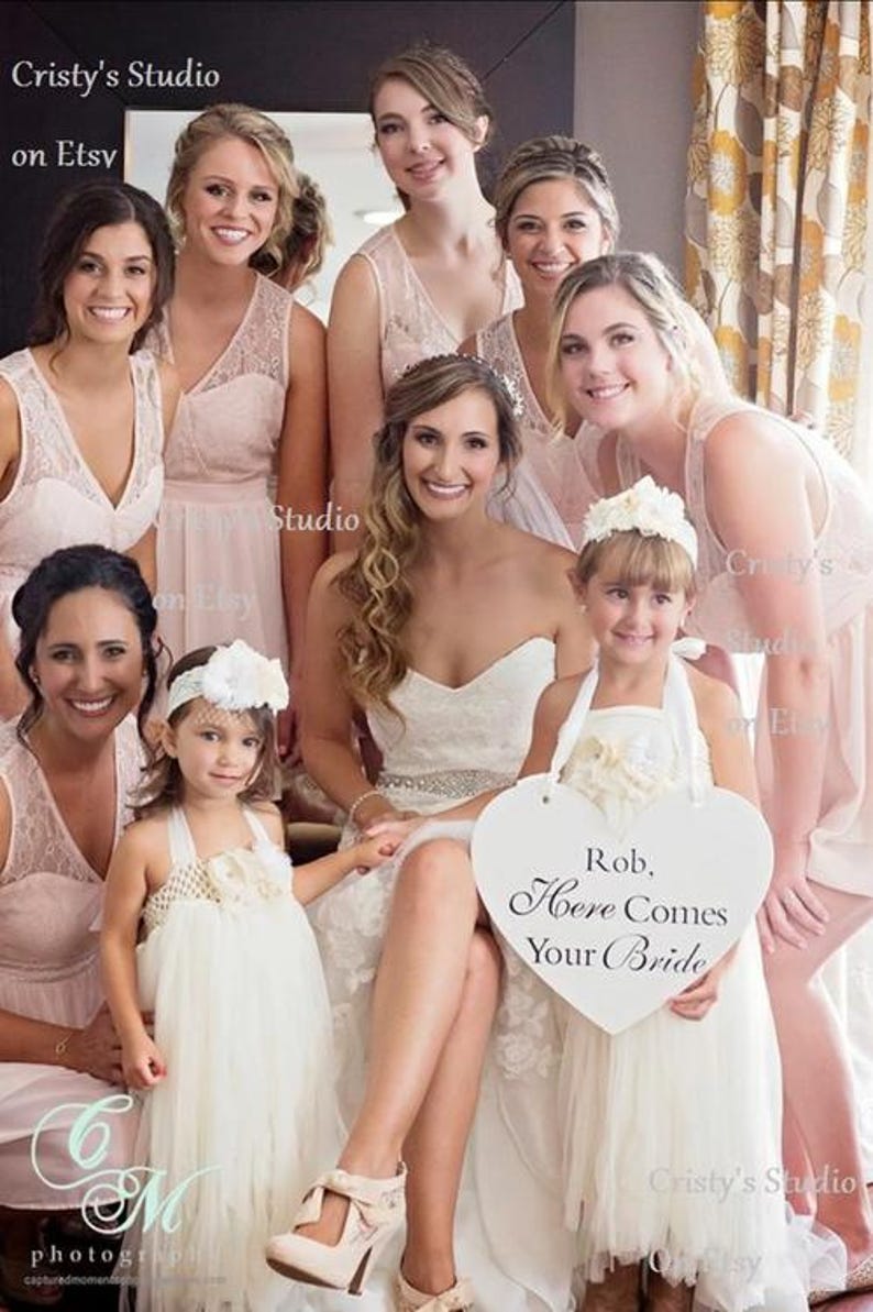 May include: A group of bridesmaids in pink dresses stand with the bride in a white wedding dress. A flower girl in a white tulle dress holds a heart-shaped sign that reads "Rob, Here Comes Your Bride".