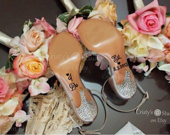 May include: A pair of bridal high heels with sparkling crystal embellishments on the heels. The soles are inscribed with "I Do" and a date. The shoes are set against a backdrop of bouquets with pink and peach roses.
