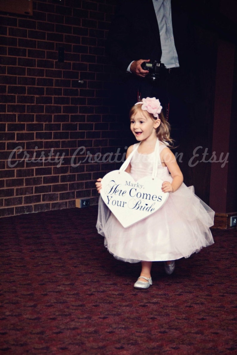 May include: A young girl in a white dress and a pink flower headband holds a white heart-shaped sign that says "Marky, Here Comes Your Bride".