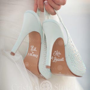 May include: A pair of light blue high-heeled wedding shoes with rhinestone embellishments. The soles are inscribed with "Est. 7.28.2013" and "Mrs. Linscott". The shoes are held up by a hand, with the text "Cristy's Studio" visible.