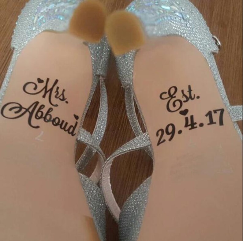 Op de afbeelding: Close-up van een paar zilveren glitter high heels. De lichtroze zolen tonen de tekst "Mrs. Abboud" en "Est. 29.4.17" in zwart script. De hakken hebben een bandjesontwerp.