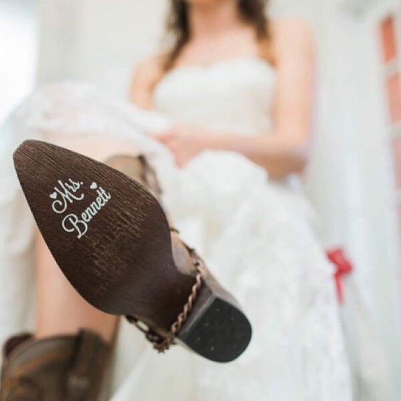 Op de afbeelding: Bruine leren cowboylaars met de tekst "Mrs. Bennett" in witte letters op de zool. De laars heeft een decoratieve ketting en een zwarte hak. Op de achtergrond een wazige bruid in een witte trouwjurk.