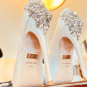 Op de afbeelding: Ivoor satijnen pumps met kristallen versieringen op de hiel. De schoenen hebben een spitse neus en een stilettohak. De woorden "Mrs. Serrano" en "Est. 4.23.22" staan op de binnenzolen. Maat 7M.