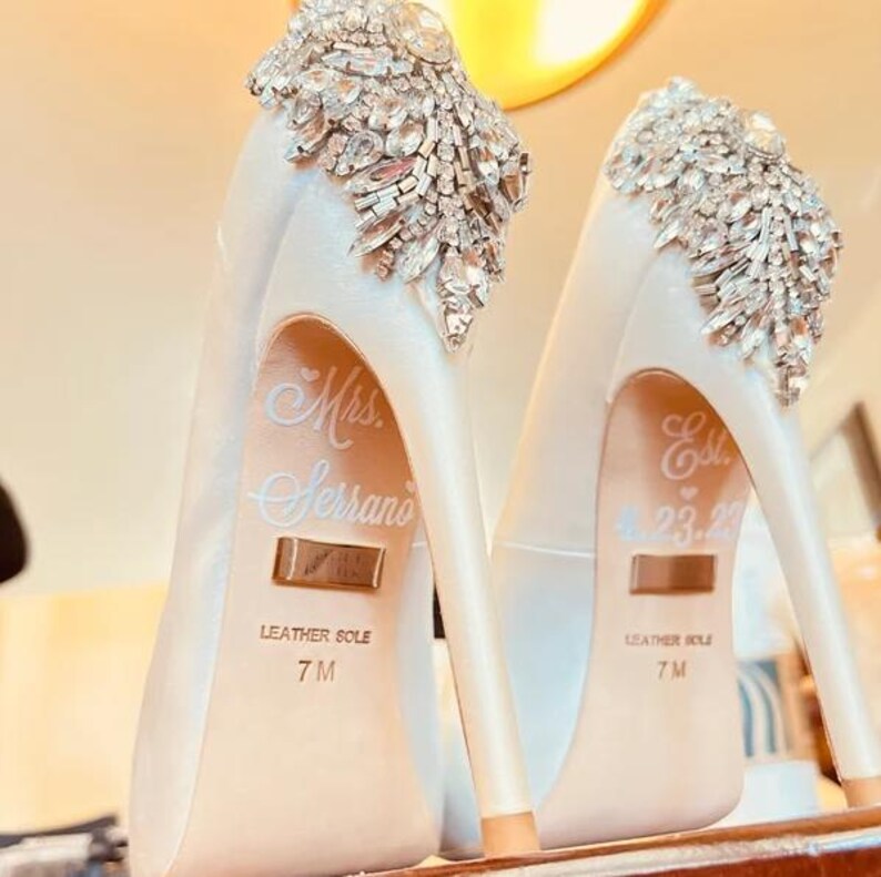 Op de afbeelding: Ivoor satijnen pumps met kristallen versieringen op de hiel. De schoenen hebben een spitse neus en een stilettohak. De woorden "Mrs. Serrano" en "Est. 4.23.22" staan op de binnenzolen. Maat 7M.