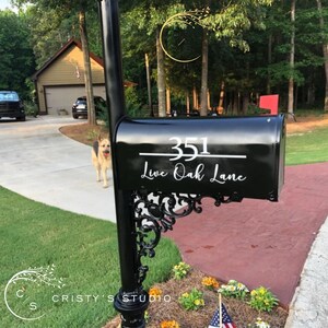 May include: Black mailbox with the address "351 Live Oak Lane" in white script. The mailbox is mounted on a black post with decorative scrollwork. A red flag is raised. A dog is in the background.
