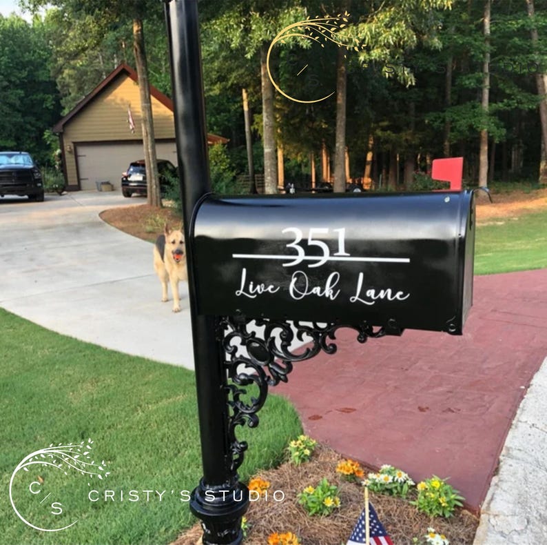 May include: Black mailbox with the address "351 Live Oak Lane" in white script. The mailbox is mounted on a black post with decorative scrollwork. A red flag is raised. A dog is in the background.