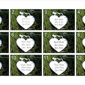 May include: Twelve white heart-shaped signs with black text that reads "Uncle Mark, Here Comes Your Bride". The signs are arranged in a grid of four rows and three columns. The signs are displayed on a green grassy background.