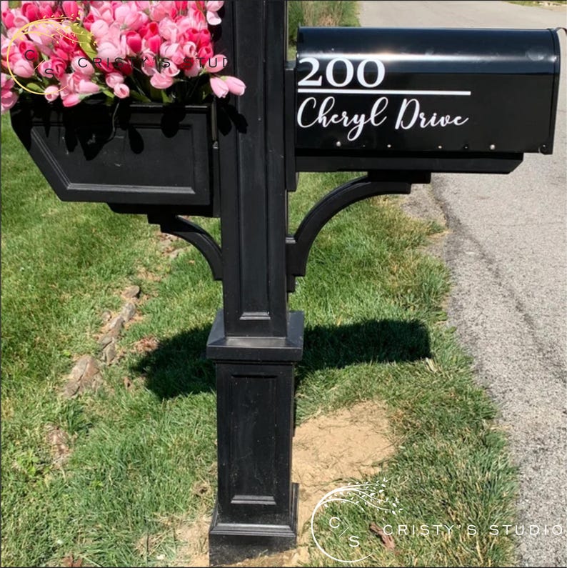 May include: Black mailbox with the address "200" and "Cheryl Drive" in white lettering. A black planter box filled with pink flowers is attached to the post. The mailbox is on a green lawn.