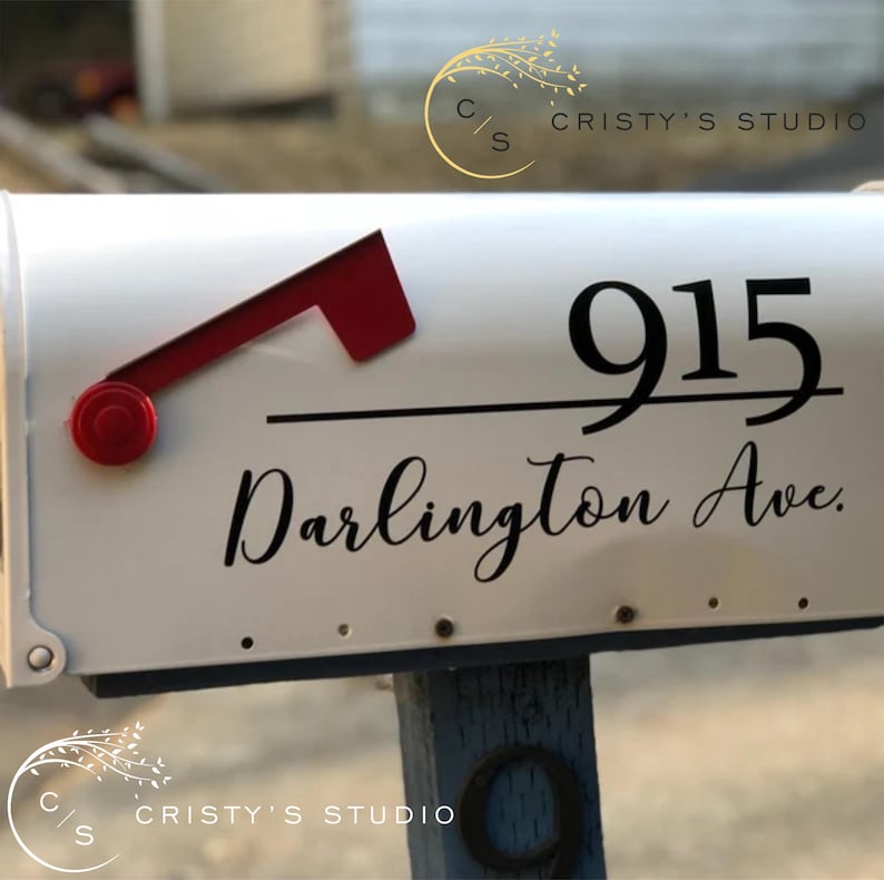 May include: White mailbox with black lettering displaying the address "915 Darlington Ave." A red flag is raised, indicating mail. The mailbox is mounted on a wooden post. The logo "Cristy's Studio" is visible.