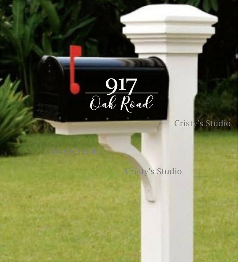 May include: A black mailbox with the address "917 Oak Road" in white script. The mailbox has a red flag and is mounted on a white post. The background includes green grass and foliage.
