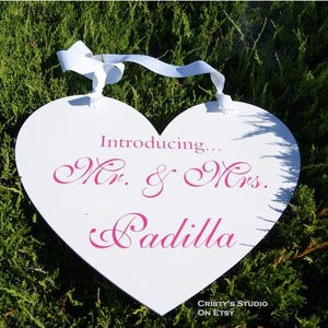 May include: White heart-shaped sign with a pink ribbon hanging from the top. The sign says "Introducing... Mr. & Mrs. Padilla" in pink cursive lettering.