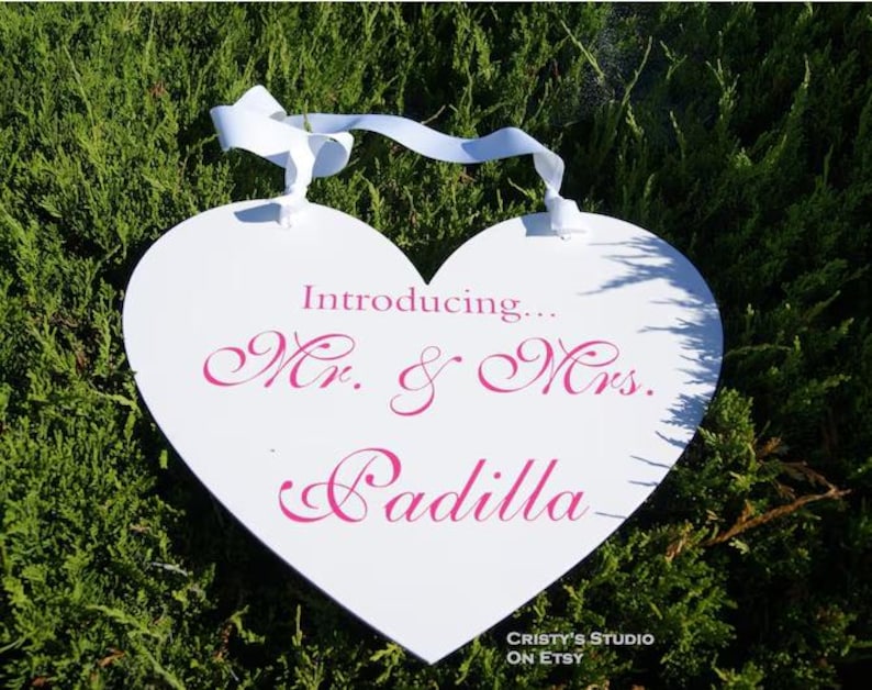 May include: White heart-shaped sign with a pink ribbon hanging from the top. The sign says "Introducing... Mr. & Mrs. Padilla" in pink cursive lettering.
