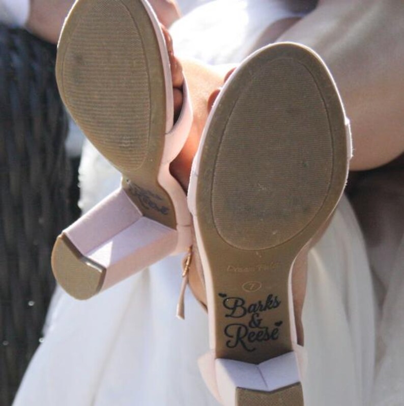 Op de afbeelding: Zachtroze sandalen met hak en blokhak. De zolen zijn lichtbruin met de tekst "Barks & Reese" in zwart schrift. De sandalen worden omhoog gehouden, waarbij de zolen zichtbaar zijn.