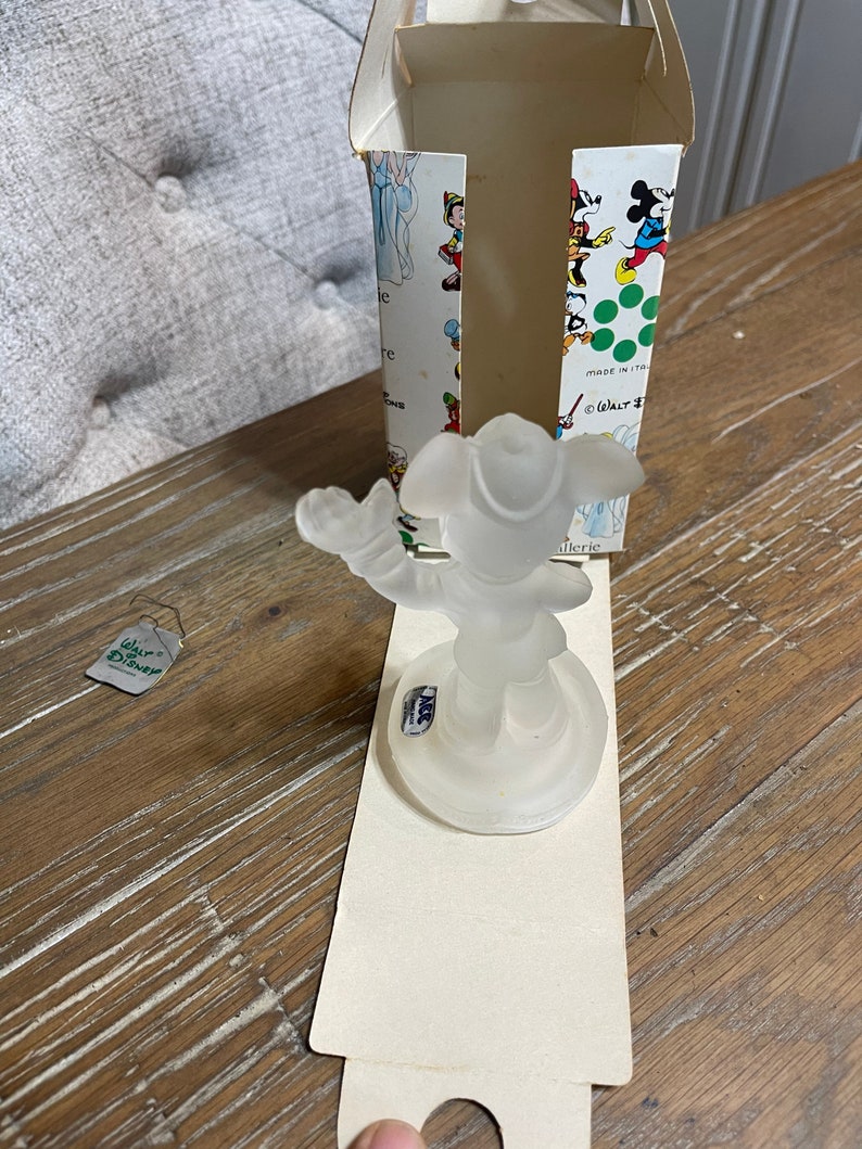 May include: A clear glass figurine of Mickey Mouse wearing a pirate hat and holding a sword. The figurine is on a clear glass base and is in a white box with a green and white pattern. The box has the text "Walt Disney" and "Made in Italy" on it.