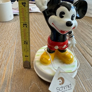 May include: A ceramic Mickey Mouse figurine with a red and white outfit, yellow shoes, and a white glove. The figurine is on a white base with green grass and is holding a string. The figurine is made by Schmid, The Creative Hand.