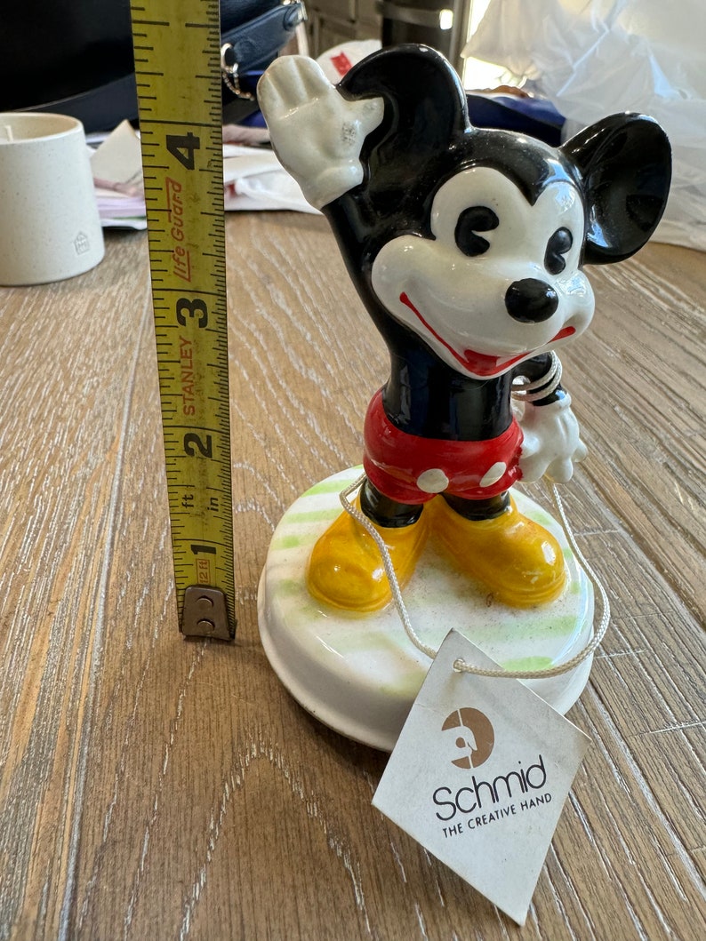 May include: A ceramic Mickey Mouse figurine with a red and white outfit, yellow shoes, and a white glove. The figurine is on a white base with green grass and is holding a string. The figurine is made by Schmid, The Creative Hand.