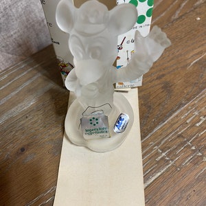 May include: A clear glass figurine of Mickey Mouse holding a musical instrument. The figurine is on a white base with a green label that reads "Imperatore oggettistica" and a blue label that reads "ACC Made in Italy".