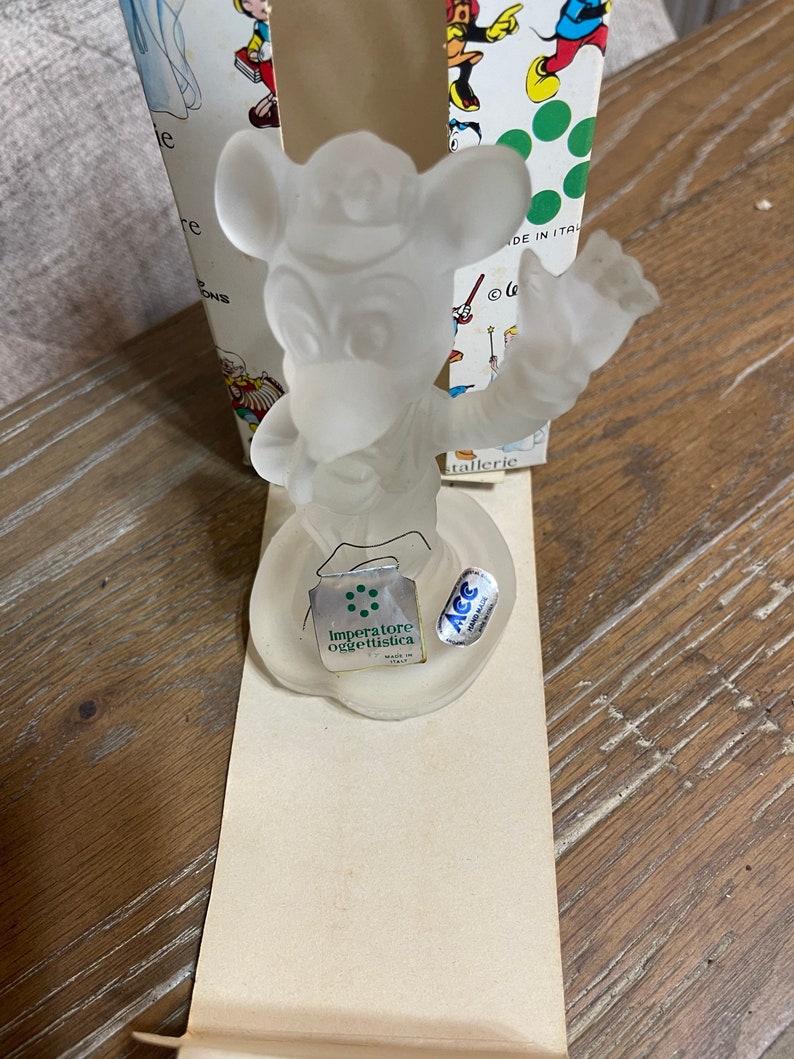 May include: A clear glass figurine of Mickey Mouse holding a musical instrument. The figurine is on a white base with a green label that reads "Imperatore oggettistica" and a blue label that reads "ACC Made in Italy".