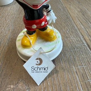 May include: A vintage porcelain Mickey Mouse figurine with a red and black outfit, yellow shoes, and a white glove. The figurine is holding a string and is on a green and white base. The base has a label that reads "Schmid The Creative Hand".