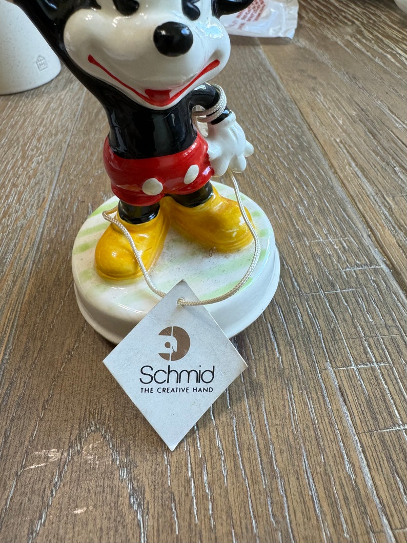 May include: A vintage porcelain Mickey Mouse figurine with a red and black outfit, yellow shoes, and a white glove. The figurine is holding a string and is on a green and white base. The base has a label that reads "Schmid The Creative Hand".