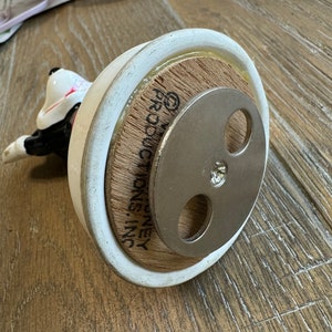 May include: White ceramic figurine of a dog with black markings. The figurine is on a wooden base with the text "G.P. MONEY INC." printed on it. The base has a metal disc with three holes on the bottom.