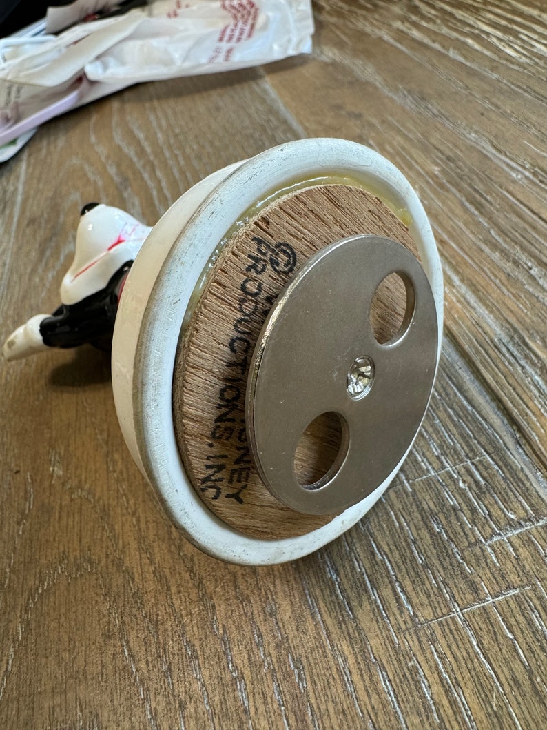 May include: White ceramic figurine of a dog with black markings. The figurine is on a wooden base with the text "G.P. MONEY INC." printed on it. The base has a metal disc with three holes on the bottom.