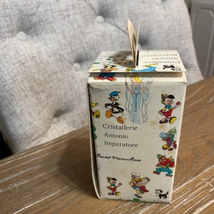 May include: A white cardboard box with a handle. The box has a colorful illustration of Disney characters, including Mickey Mouse, Donald Duck, and Pinocchio. The text on the box reads "Cristallerie Antonio Imperatore" and "Disney Productions".