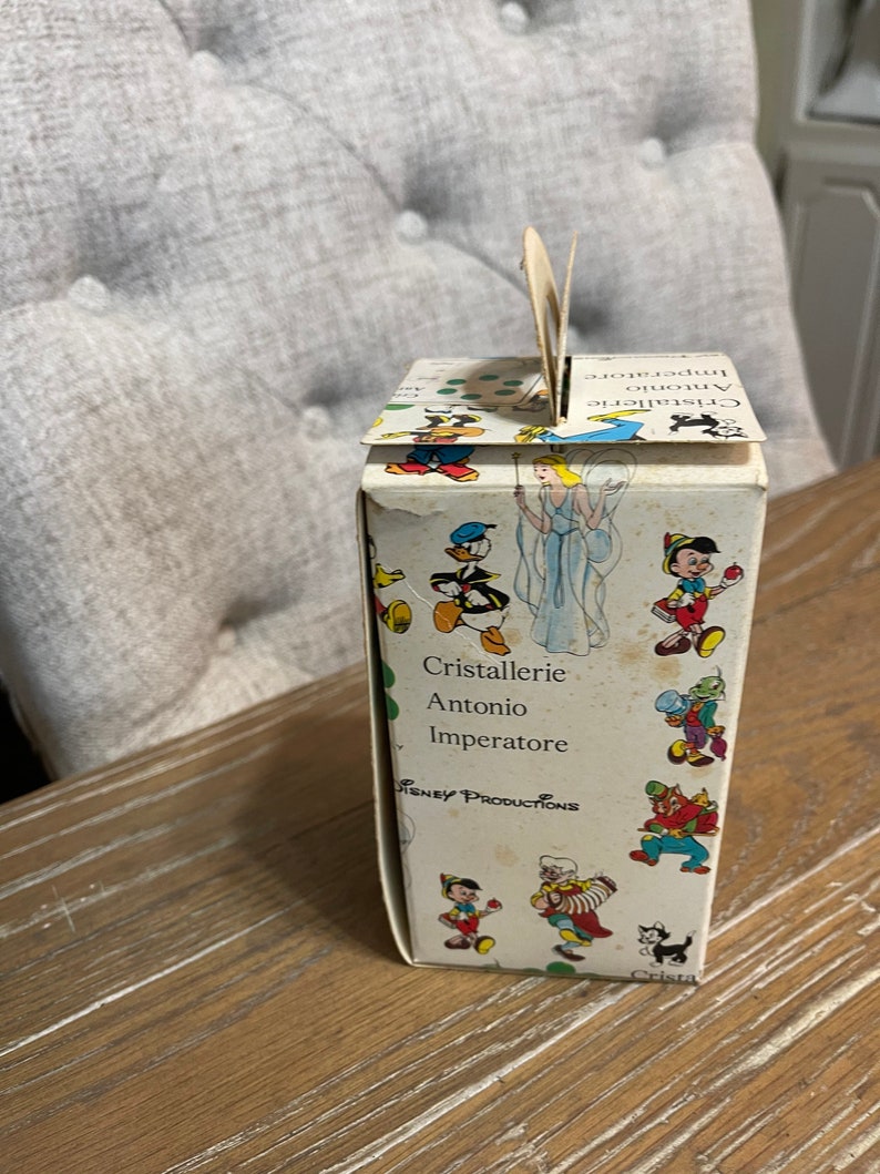 May include: A white cardboard box with a handle. The box has a colorful illustration of Disney characters, including Mickey Mouse, Donald Duck, and Pinocchio. The text on the box reads "Cristallerie Antonio Imperatore" and "Disney Productions".