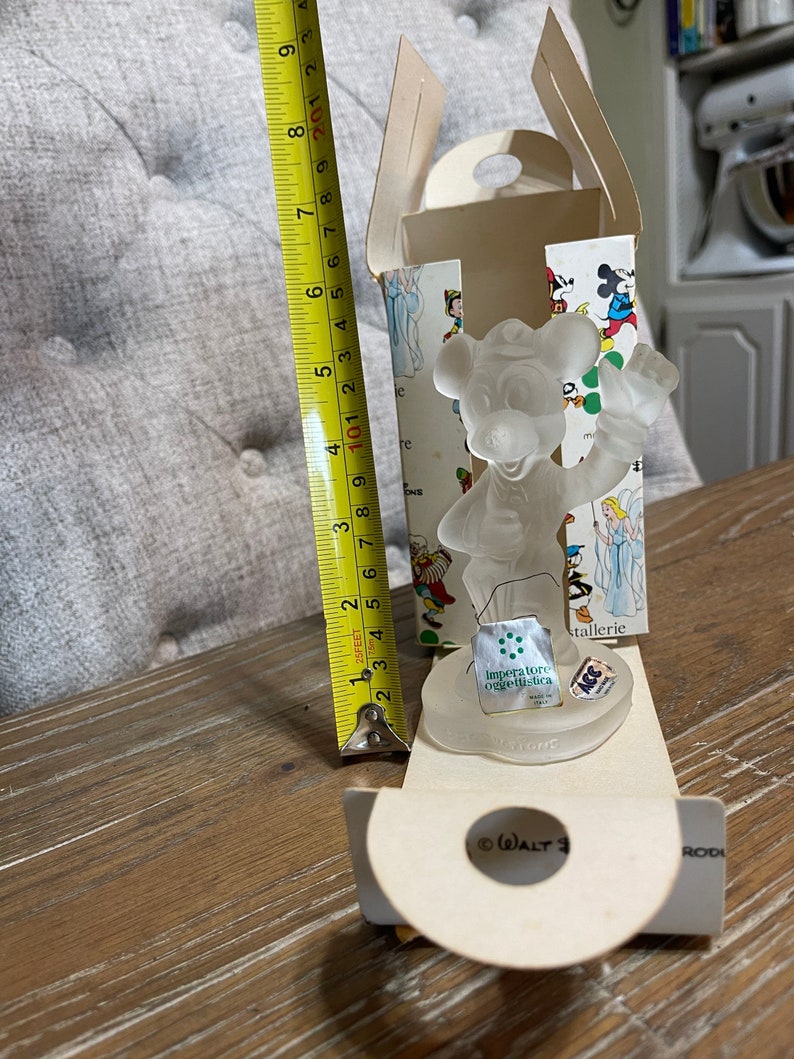 May include: A clear glass figurine of Mickey Mouse holding a musical instrument. The figurine is in a white box with a colorful illustration of Mickey Mouse and other Disney characters. The box also includes the text "Imperatore oggettistica" and "ACC".