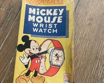 Könnte beinhalten: Vintage Ingersoll Mickey Mouse Armbanduhr-Box mit einem Cartoon-Bild von Mickey Mouse, der die Uhr hält. Die Box ist gelb mit blauen und roten Akzenten. Der Text auf der Box lautet "Ingersoll Mickey Mouse Wrist Watch" und "US Time".